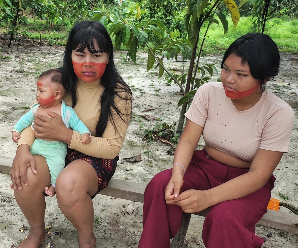 Woman holding a baby seated beside another woman in a wooded area